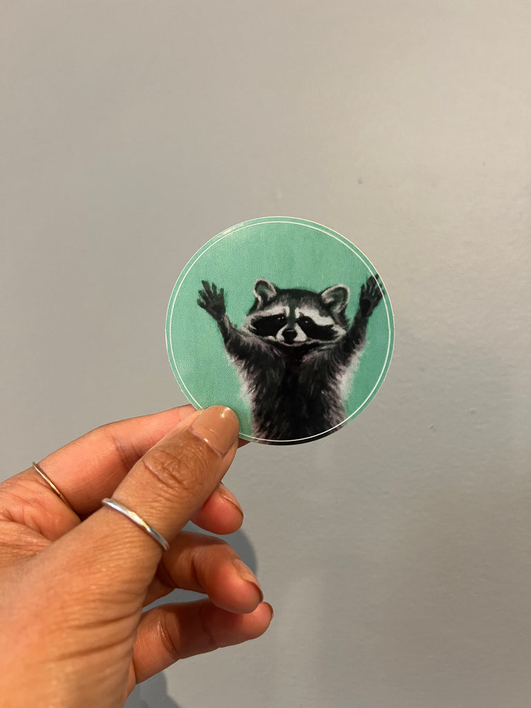 Circular sticker with a raccoon design held by a hand against a neutral background